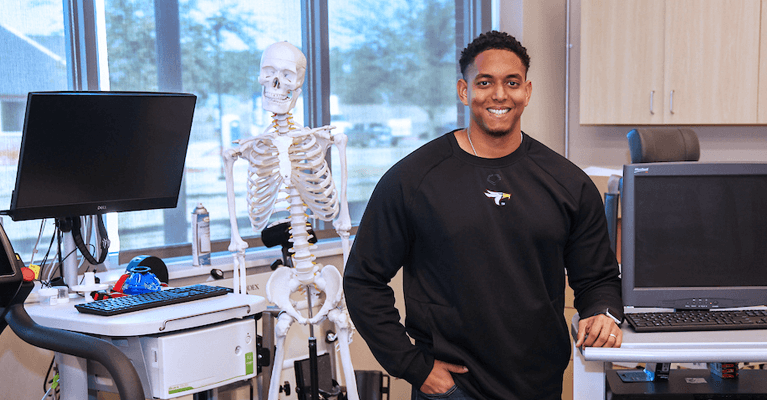 Smiling man standing in a medical imaging classroom next to a full-size skeleton model, with computer workstations and large windows in the background.